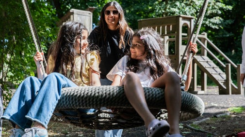 Two children are sitting sharing a basket swing. There is an adult standing behind them and behind them there is a wooden climbing frame. The sun is shining brightly through the trees